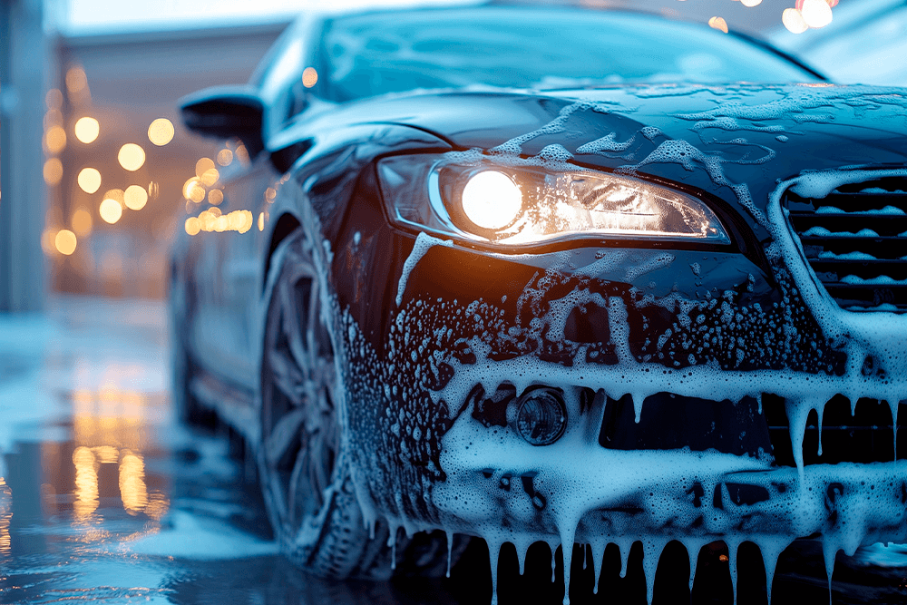 Close-up of a dark-colored car covered in thick soap suds during a car wash, with its headlight on and water reflecting on the wet ground. Blurred background with warm lights visible.